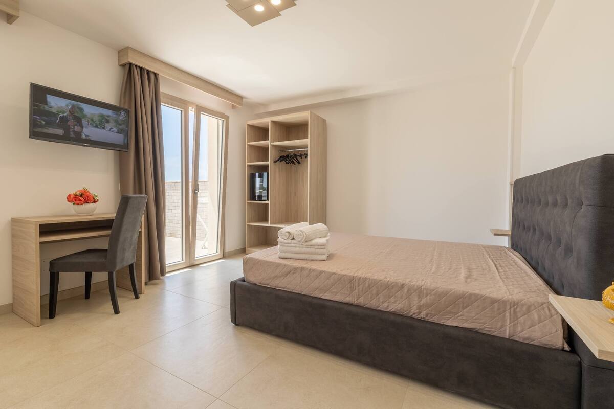 A modern hotel room with a bed, a desk, a chair, a TV, and a wardrobe.