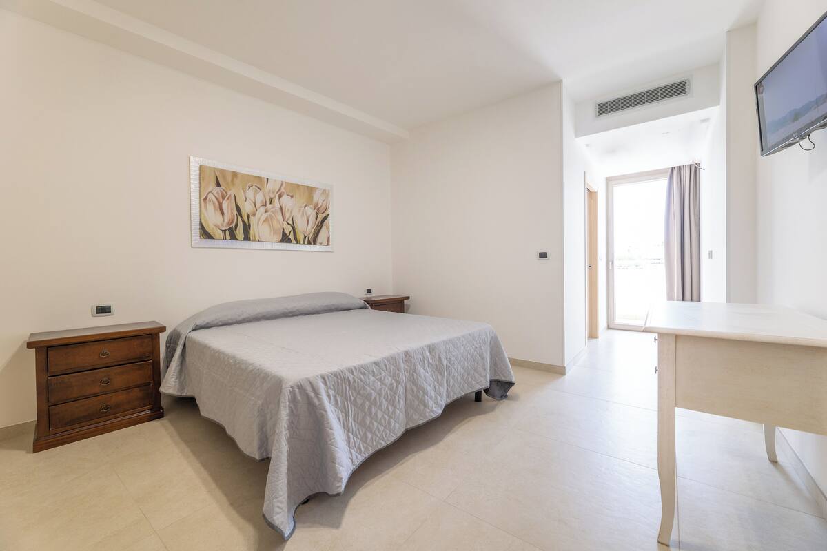 A bedroom with a bed, a dresser, a desk, a painting, and a television.