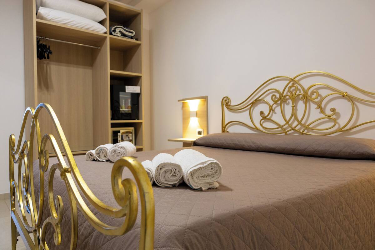 A neatly made bed with rolled towels, a bedside table with a lamp, and a wardrobe with shelves.