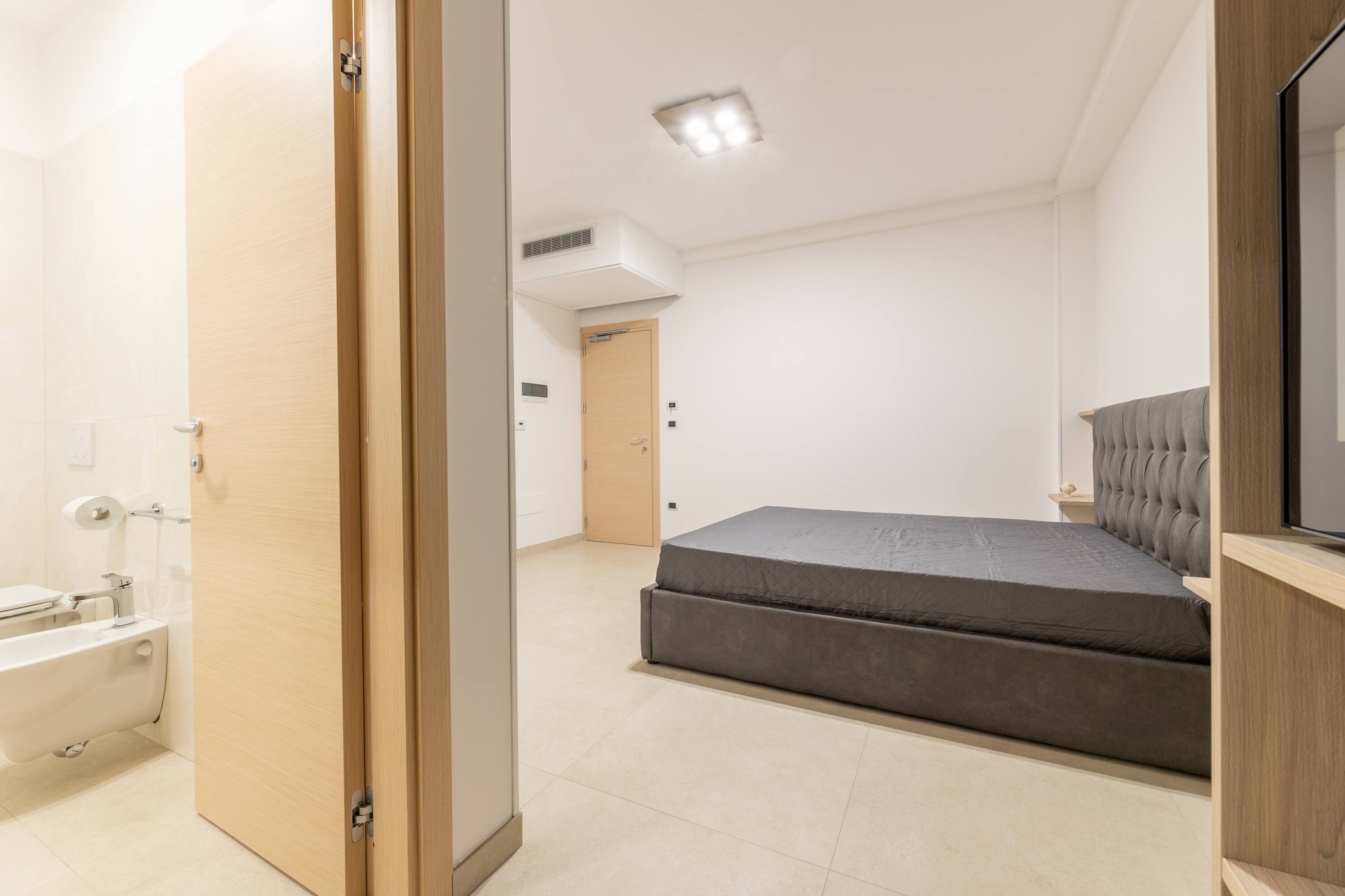 A modern bedroom with a bed, a wooden door, a bathroom with a bathtub, and a ceiling light.