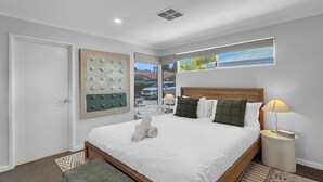 Un dormitorio ordenado con una cama grande, una mesita de noche con lámpara, una obra de arte en la pared y una ventana con vistas al exterior.