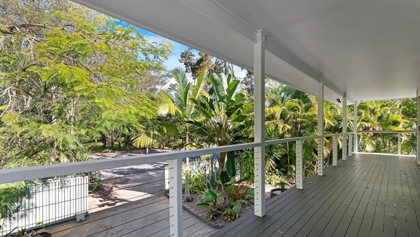 Un espacio al aire libre techado con terraza de madera, barandillas blancas y exuberante vegetación tropical.