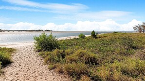 Un camino de arena que lleva a un mar tranquilo, con cielo despejado y tierra a lo lejos.