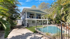 Una casa de dos pisos con balcón, rodeada por una piscina y vegetación tropical.