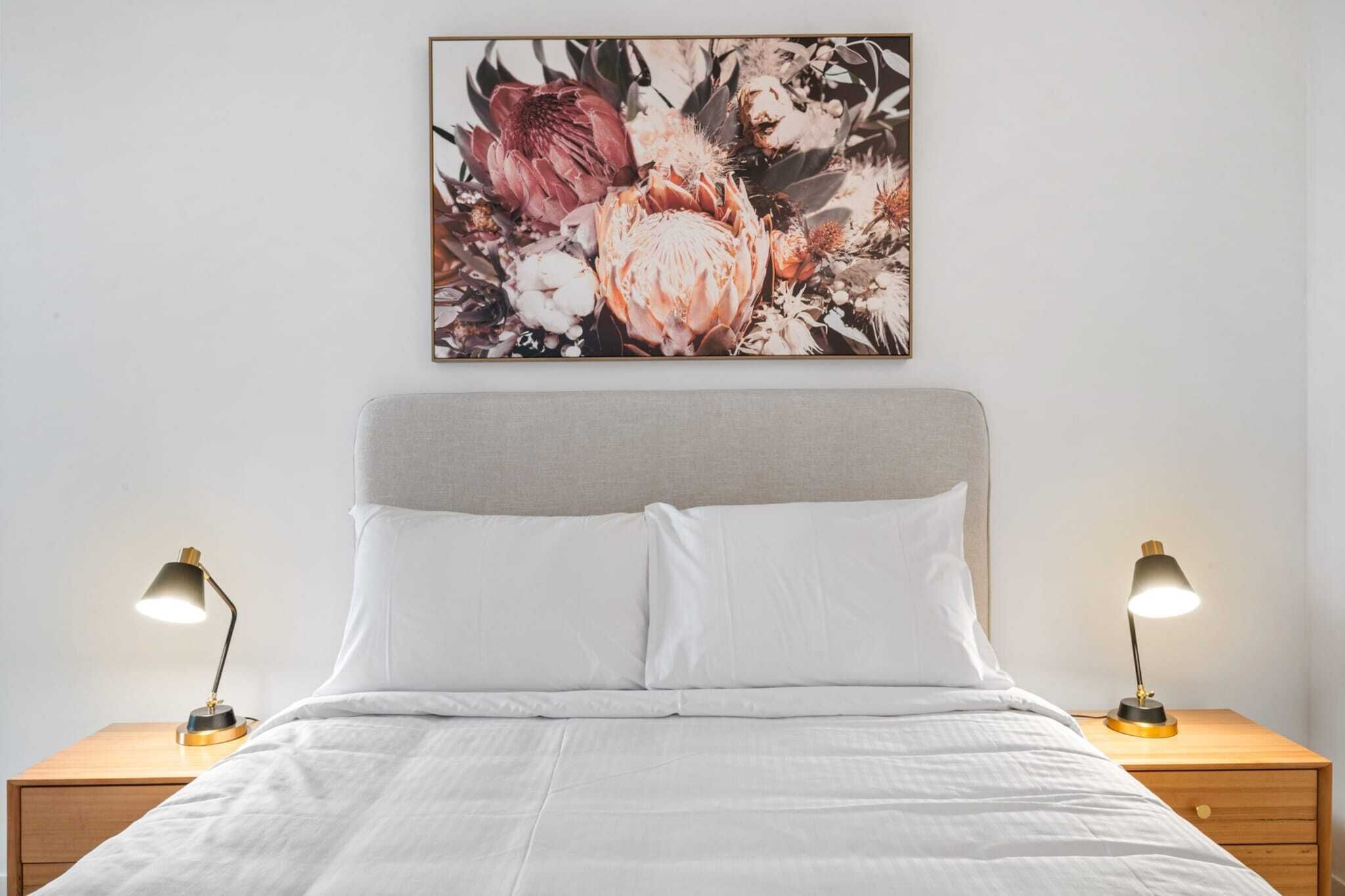 Un lit bien fait, avec du linge de lit blanc, deux tables de chevet avec des lampes et une œuvre d’art florale accrochée au mur.