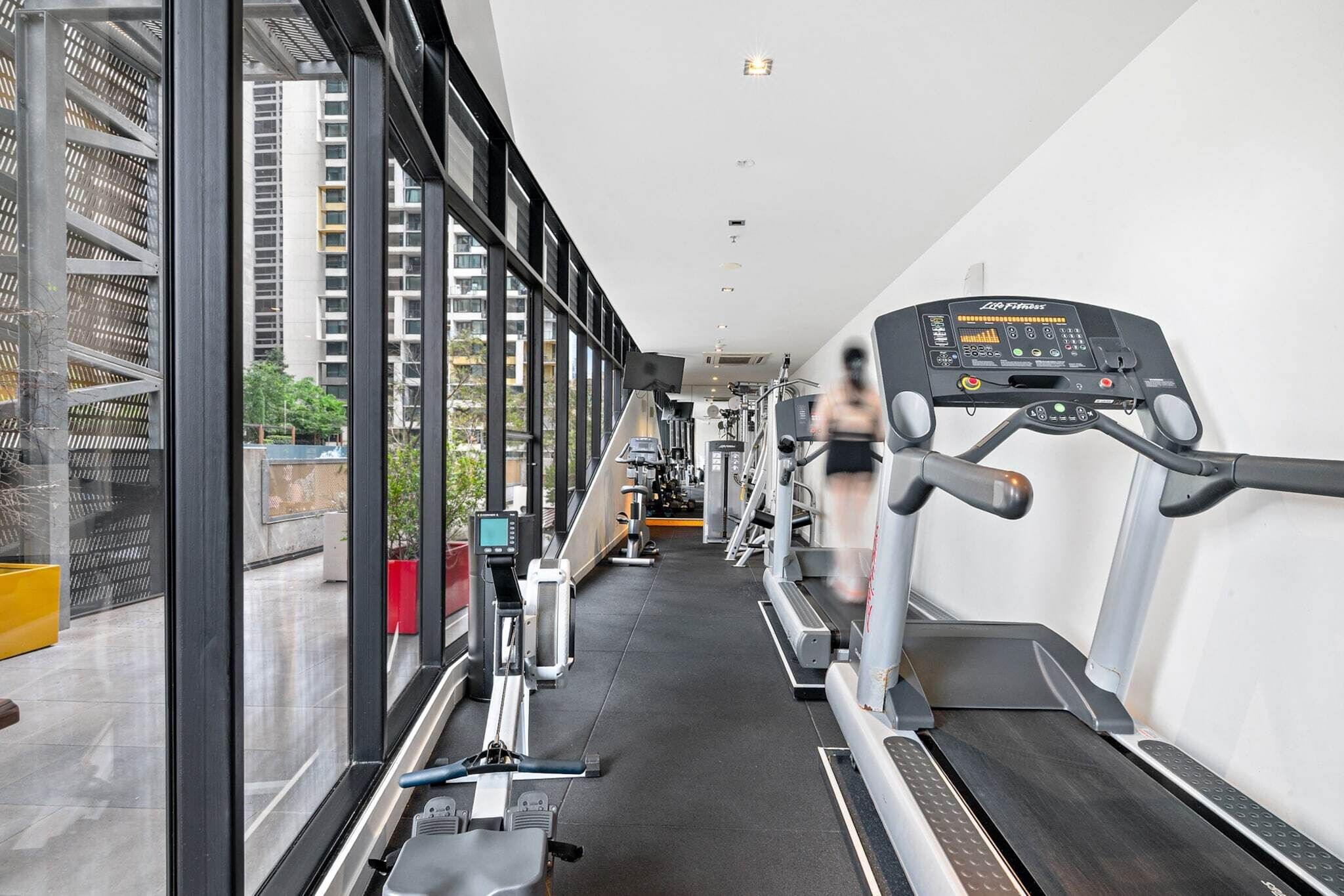 A gym with treadmills, exercise equipment, and large windows offering views of buildings and greenery.
