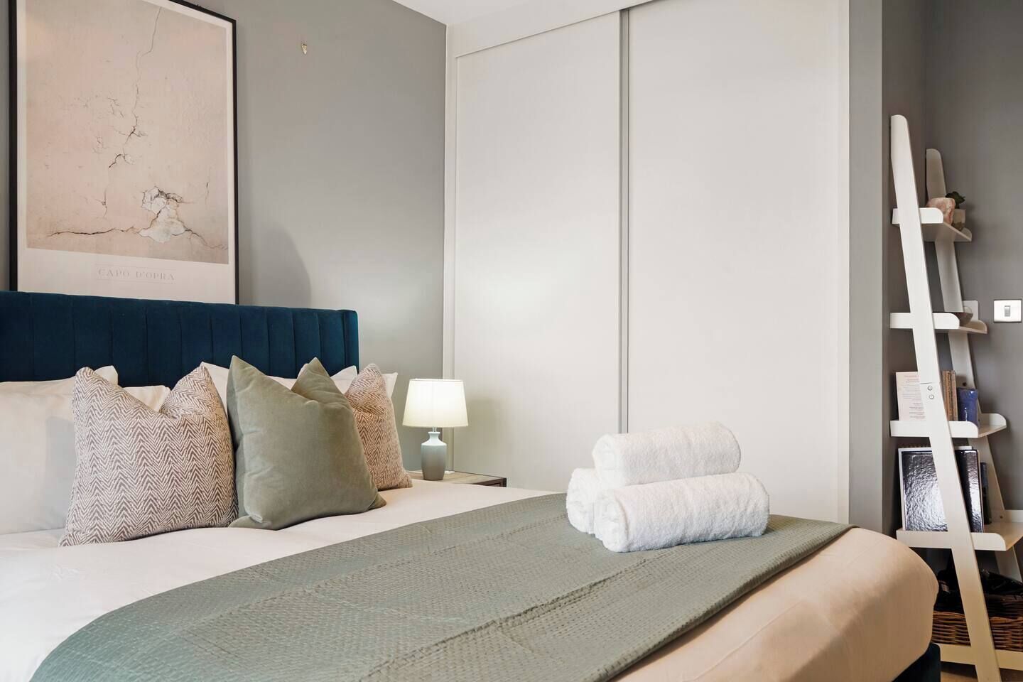 A hotel room with a bed, pillows, a bedside table with a lamp, a towel rack, and a framed picture on the wall.