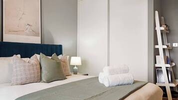 A hotel room with a bed, pillows, a bedside table with a lamp, a towel rack, and a framed picture on the wall.