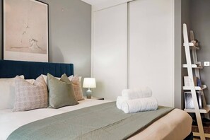 A hotel room with a bed, pillows, a bedside table with a lamp, a towel rack, and a framed picture on the wall. - Stylish 1-Bed Apt with Balcony & Canal Views (Feltham)