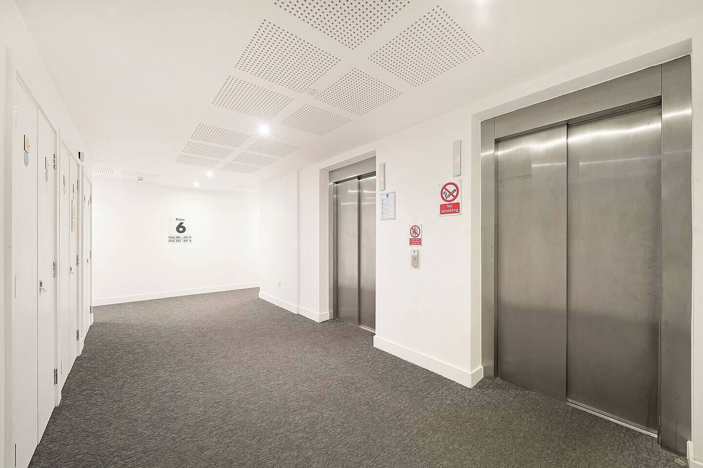 A hallway with a carpeted floor, white walls, and two elevators.