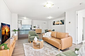 Ein modernes Wohnzimmer mit einem Ledersofa, einem grünen Sessel und einem Fernseher auf einem Holzschrank. Im Hintergrund ist eine Küche mit weißen Schränken und einem Essbereich zu sehen.