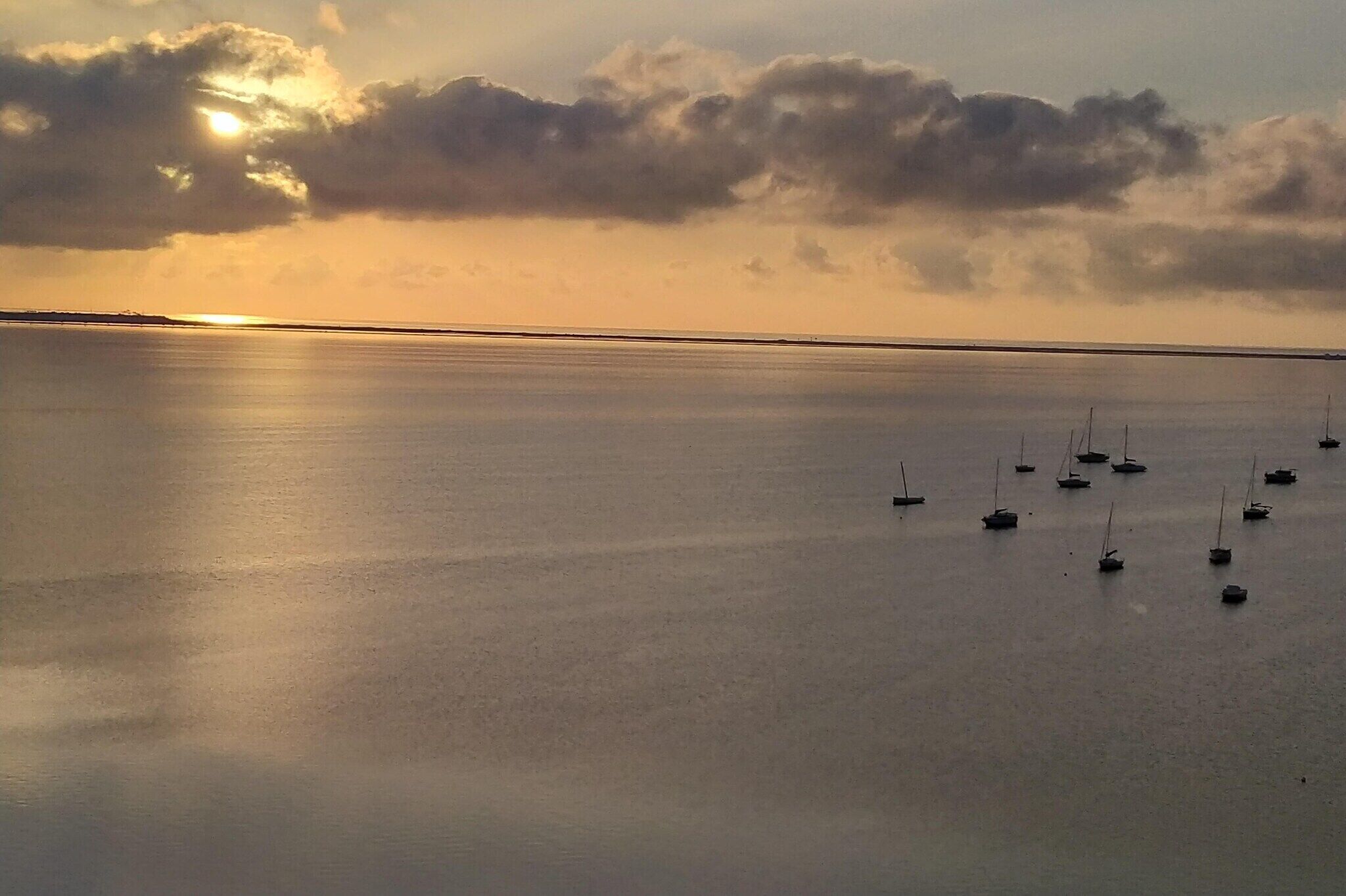 A calm body of water with several boats, a clear horizon, and a sun partially obscured by clouds.