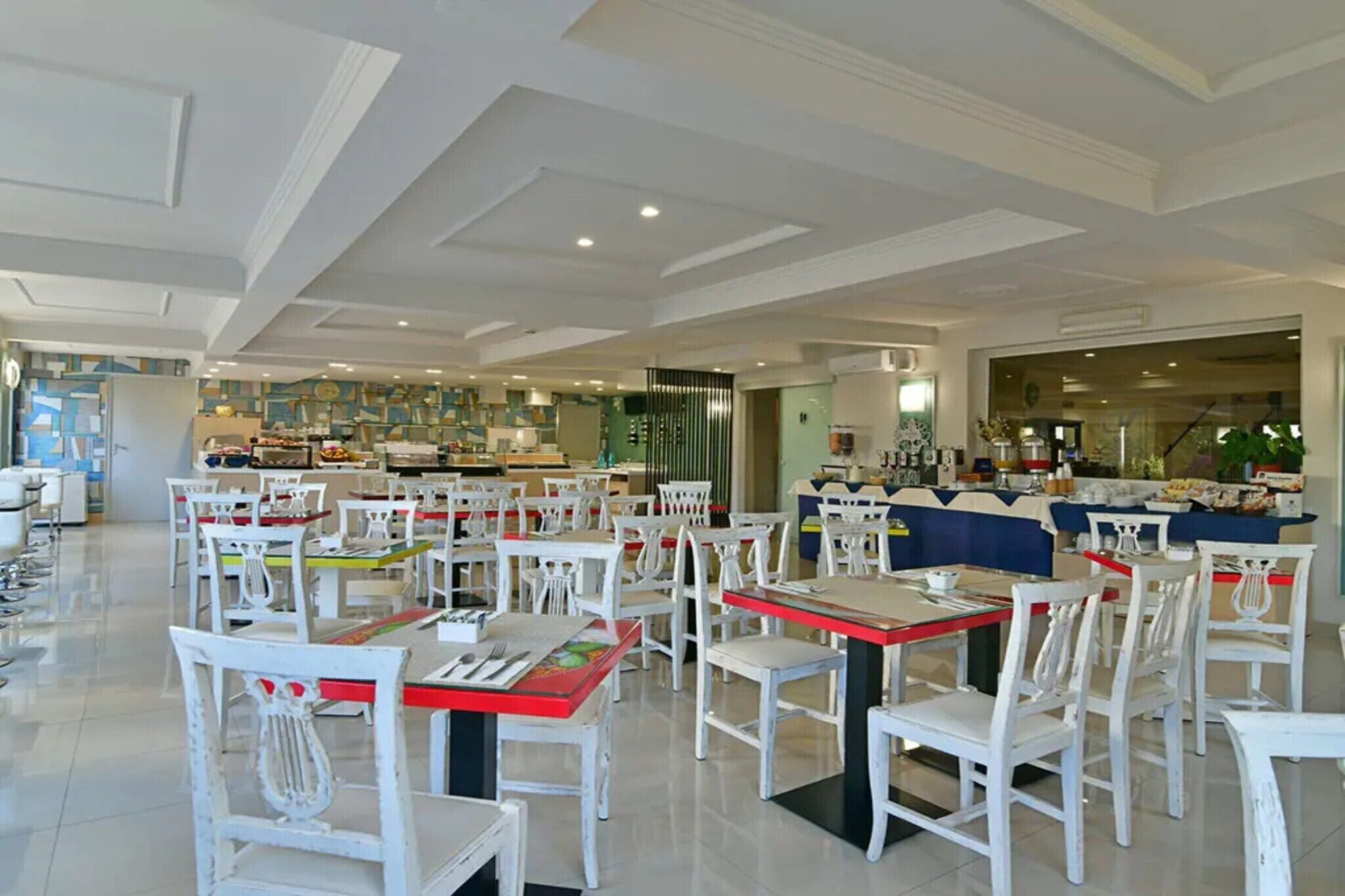 A spacious dining area with white chairs and red tables, a kitchen in the background, and a modern ceiling design.