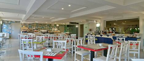 A spacious dining area with white chairs and red tables, a kitchen in the background, and a modern ceiling design.