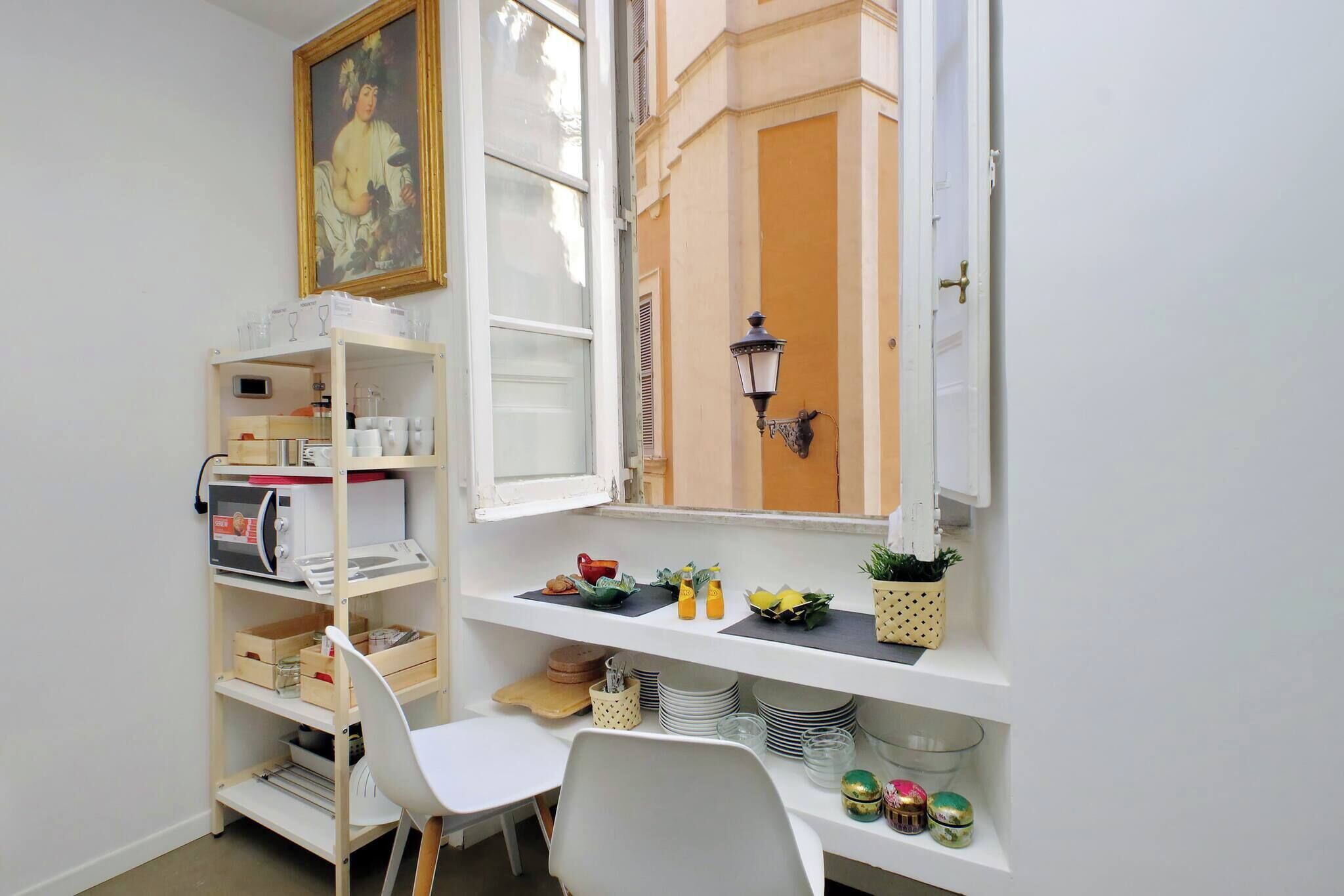 Une cuisine de taille réduite, avec des meubles blancs, une chaise blanche et une fenêtre donnant sur un immeuble.
