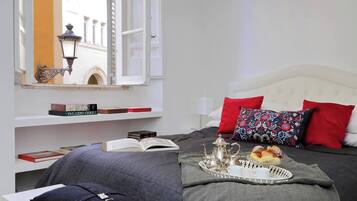 Un dormitorio ordenado con cama, almohadas, una bandeja con pasteles, un libro abierto y una ventana con vista a edificios.