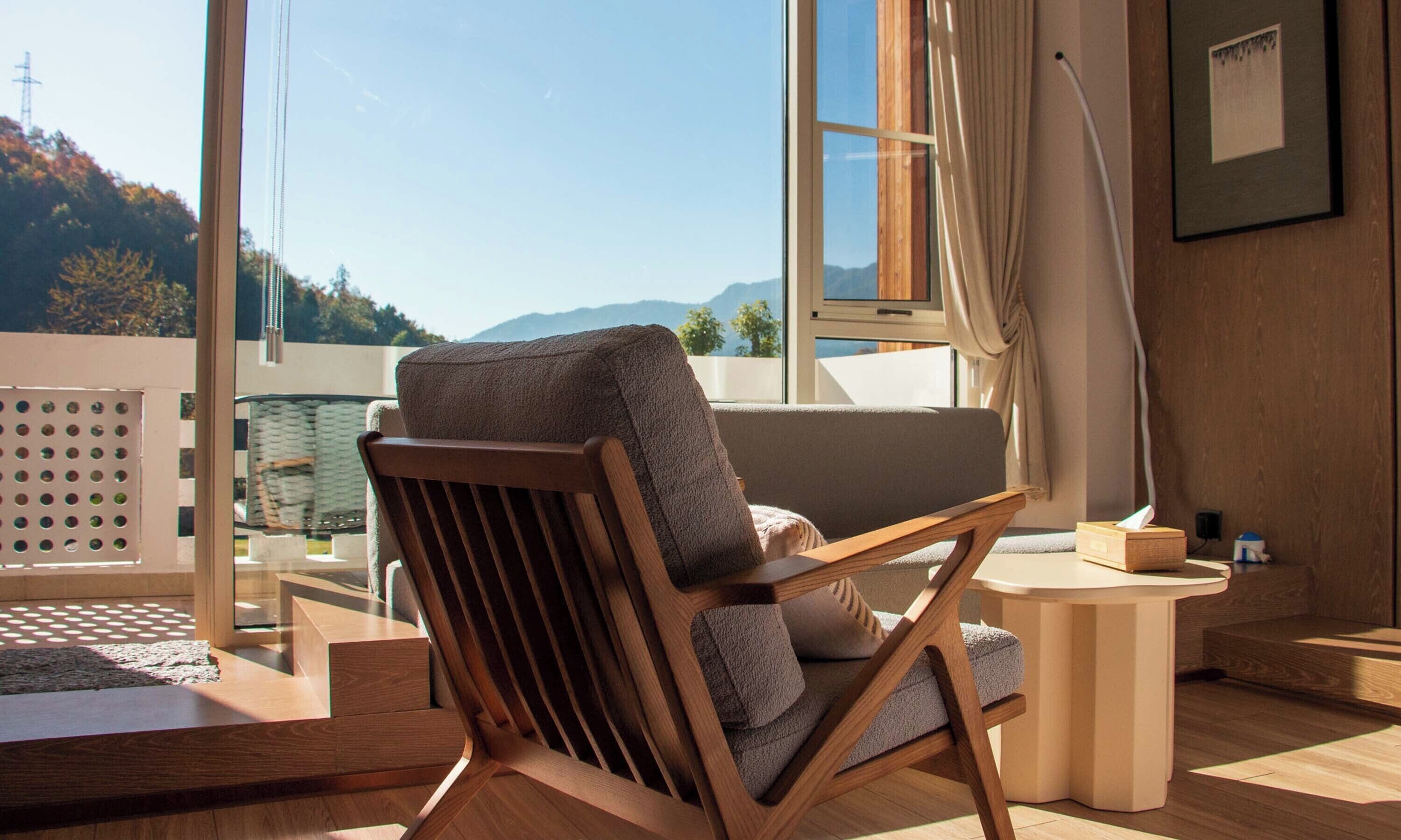 A modern room with a wooden chair, a small table, and a view of mountains through large windows.