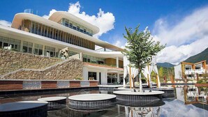 A modern hotel with a curved facade, a stone retaining wall, and a reflective pool with circular platforms. - Old River Story Art Mountain Retreat (Mianyang)