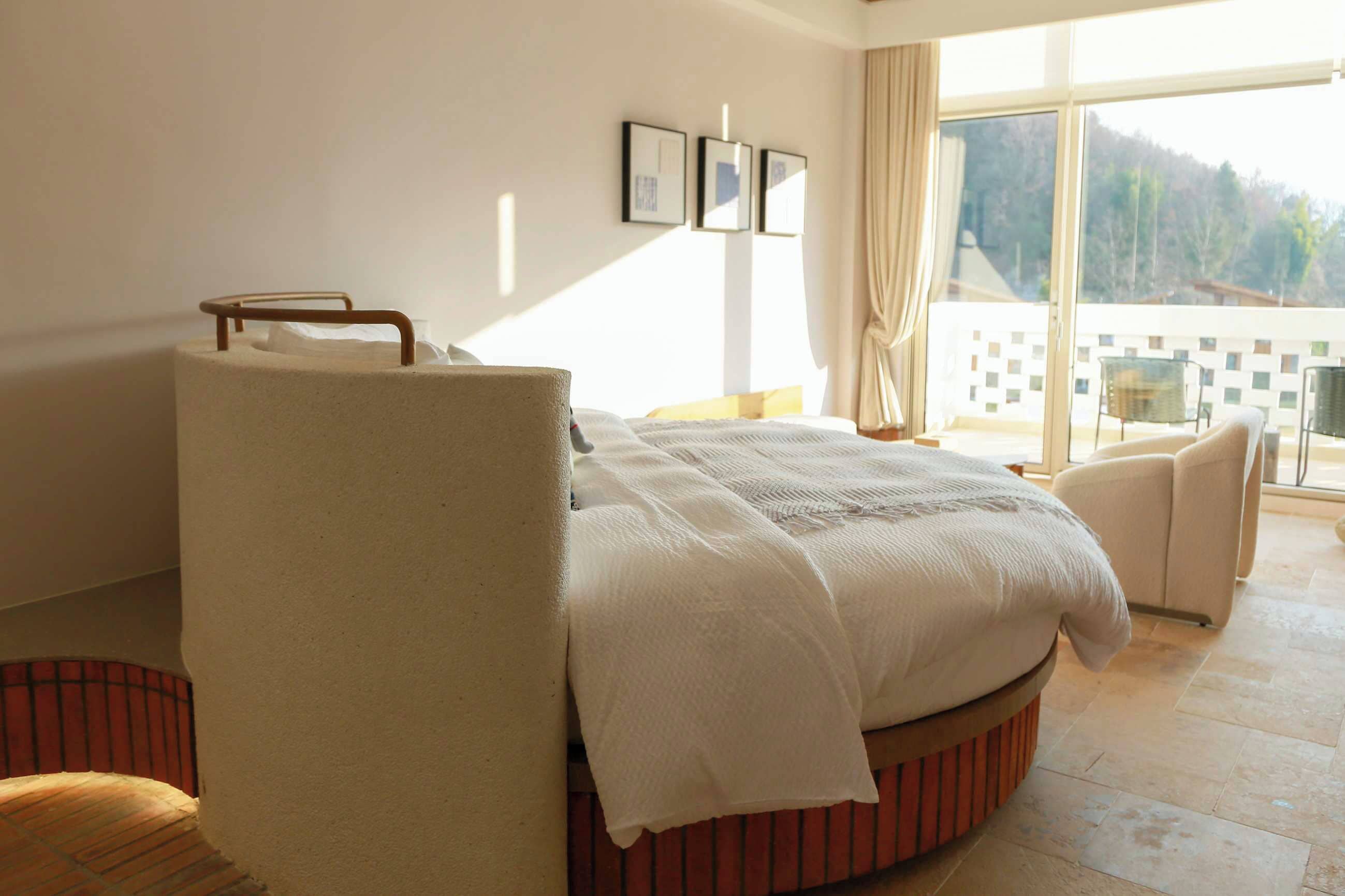 A bedroom with a bed, a chair, a large window, and a view of the outdoors.