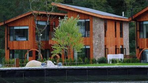A modern wooden cabin with a stone facade, situated by a calm body of water, surrounded by dense forest. - Old River Story Art Mountain Retreat (Mianyang)