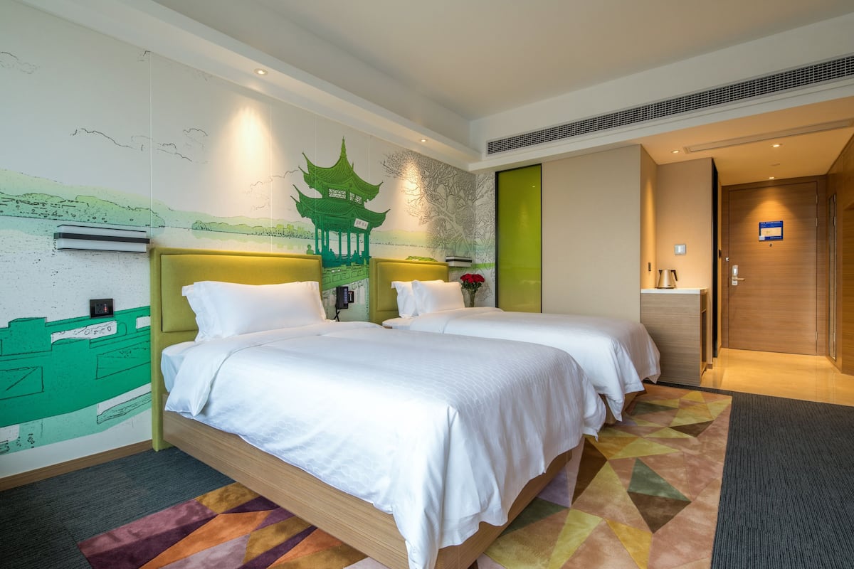 Une chambre d’hôtel avec deux lits, une fresque murale verte, une porte en bois et un petit bureau.