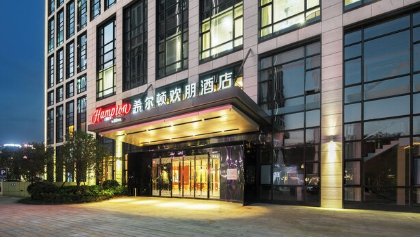 A hotel with a modern facade, large windows, and a prominent entrance with illuminated signage. - Hampton by Hilton Hangzhou Binjiang (Hangzhou)