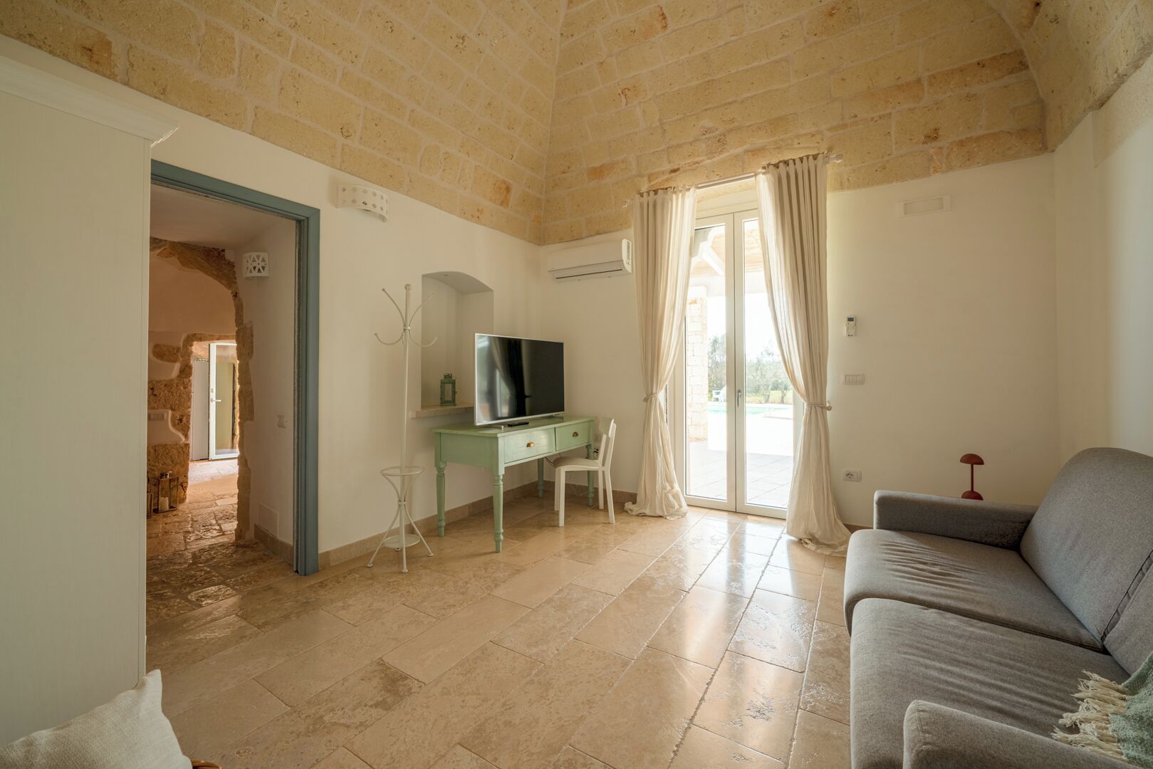 A room with a stone ceiling, a television on a green desk, a grey sofa, and a door leading to an outdoor area.