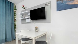 Et lille rum med et hvidt bord, to hvide stole, et ophængt tv og et billede på væggen.