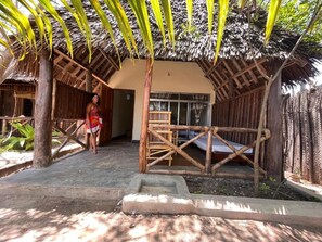 A thatched-roof structure with a bed and a small table, surrounded by wooden fences and plants. - Ushongo beach bandas (Pangani)