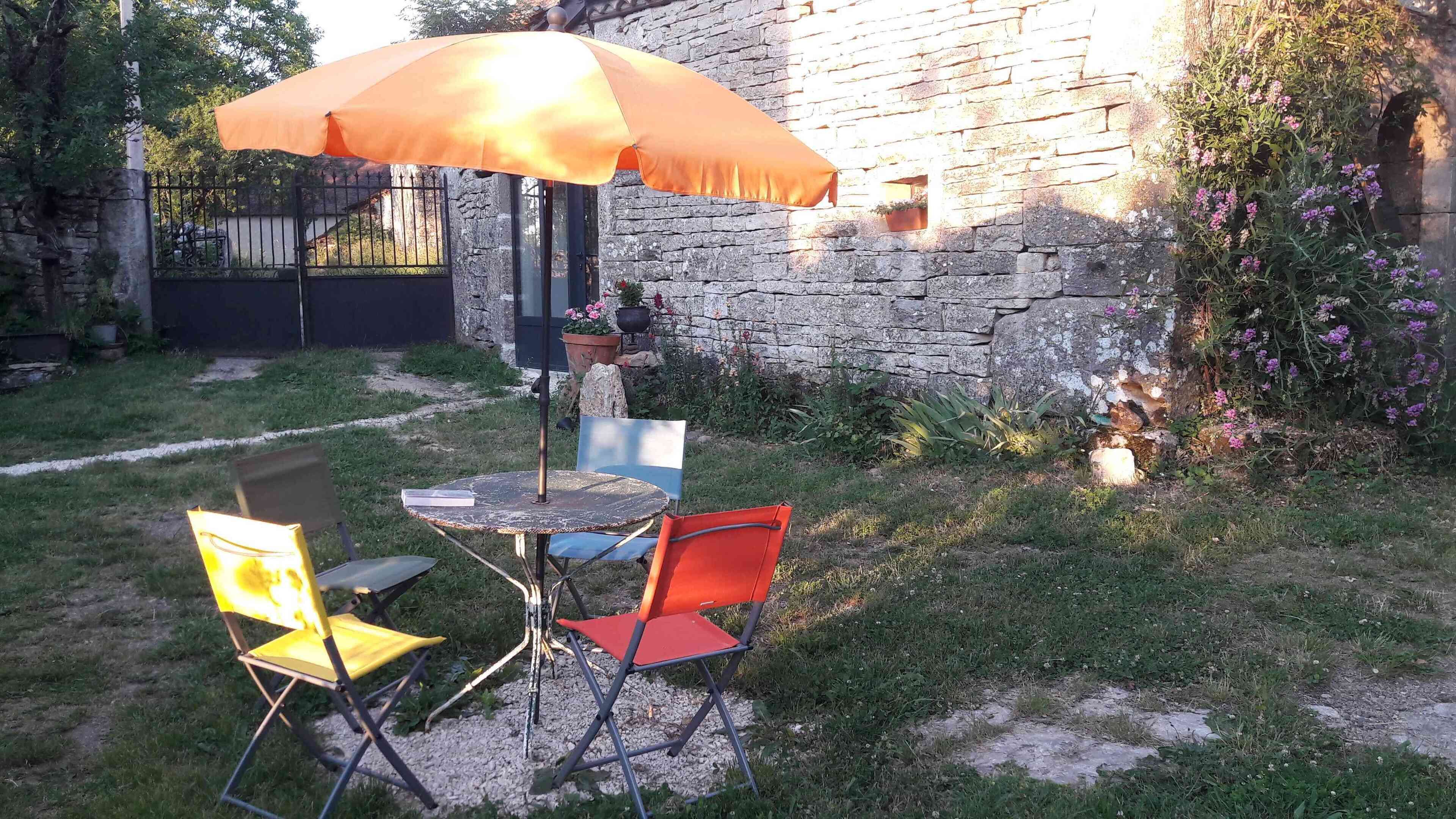 Ein Sitzbereich im Freien mit einem Tisch und zwei Stühlen, einem orangefarbenen Sonnenschirm und einer Steinmauer im Hintergrund.