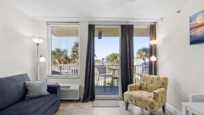 A living room with a sofa, armchair, and a table. There is a balcony with a view of palm trees and a railing. - Harbour Beach Resort 219 (Daytona Beach)