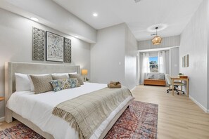 A bedroom with a large bed, a bedside table, a desk, and a chair. There is a window with curtains and a view of the outside. - 7BR 5BA w Pool, Hot Tub, Sport Court & Game Room (DeSoto)