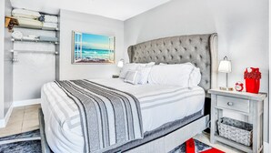 A bedroom with a large bed, a nightstand, a wall-mounted shelf with towels, and a framed picture of the ocean. - Ocean Jewels Club 104 - Beach Jewel (Daytona Beach)