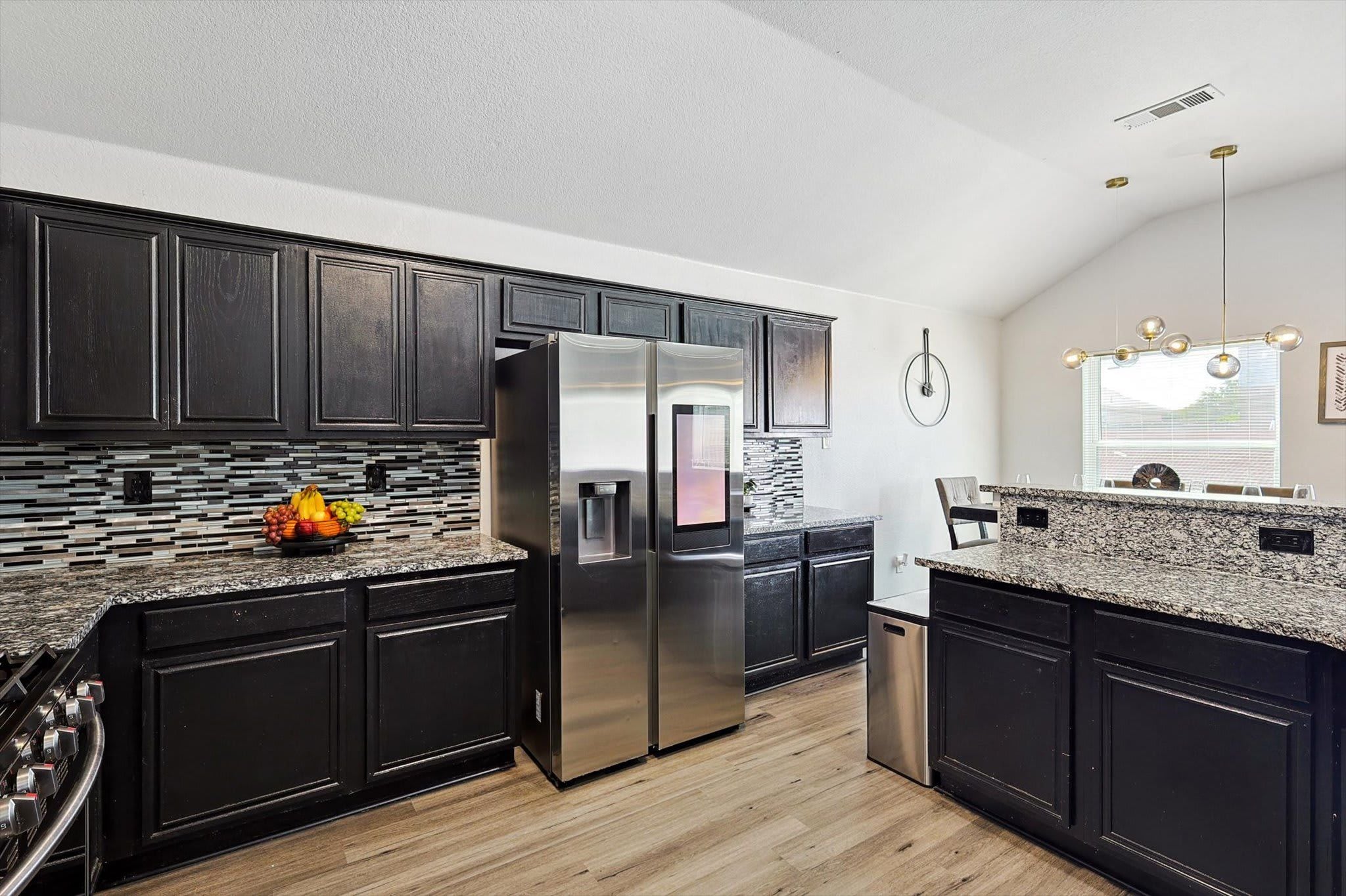 A modern kitchen with dark cabinetry, a stainless steel refrigerator, granite countertops, and a fruit bowl.