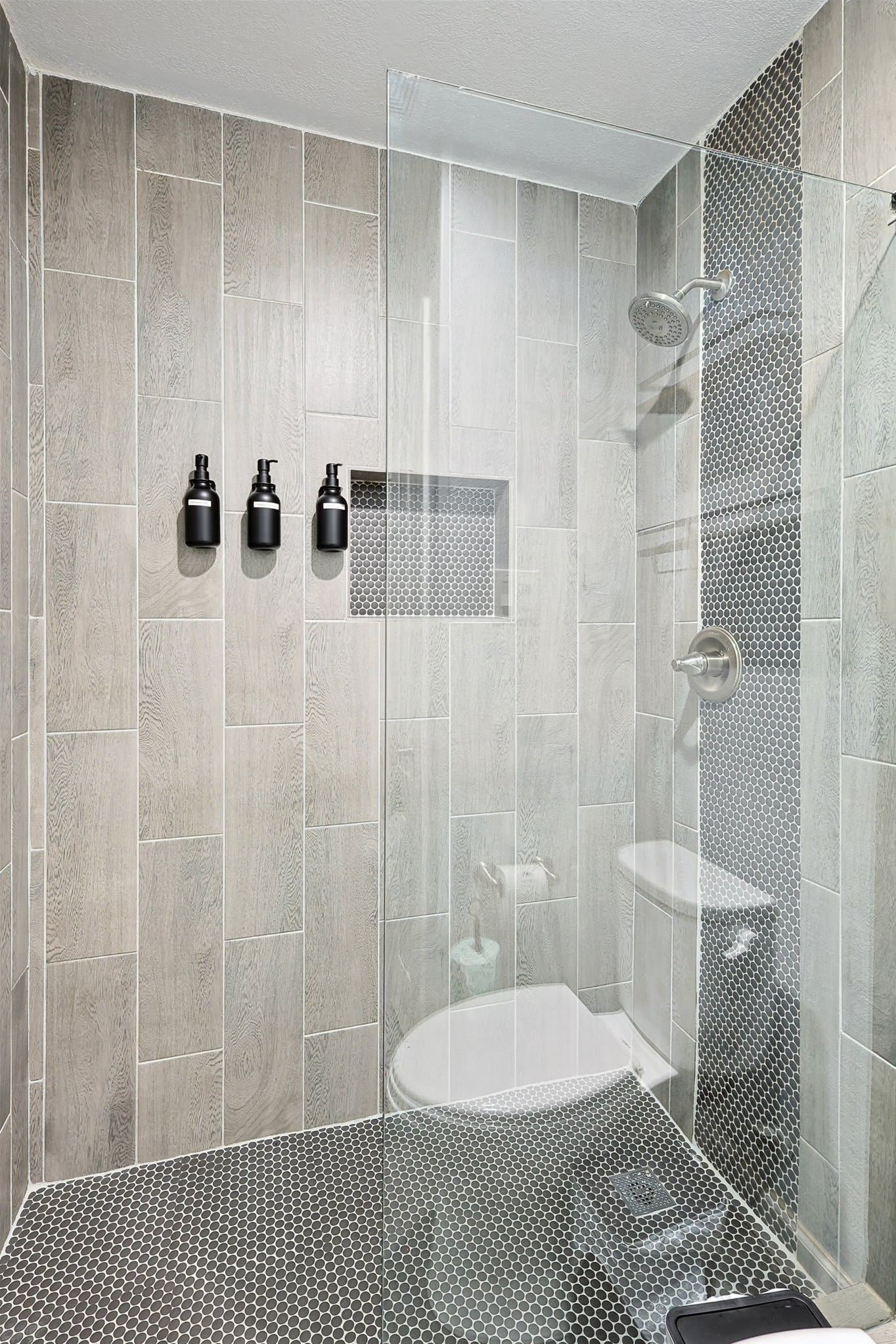 A modern shower with a glass door, a white sink, and three black dispensers on the wall.