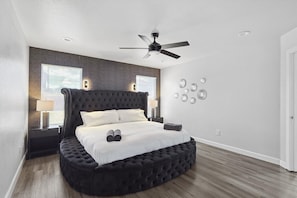 A modern bedroom with a tufted headboard, a round bed, a ceiling fan, and a brick accent wall. - 5BR Getaway in Desoto With Pool, Hot Tub & Cinema (DeSoto)
