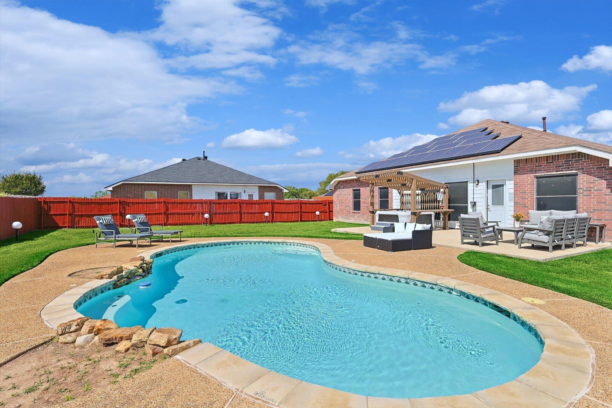 Un patio trasero con piscina, un porche con mobiliario exterior y una casa con paneles solares.