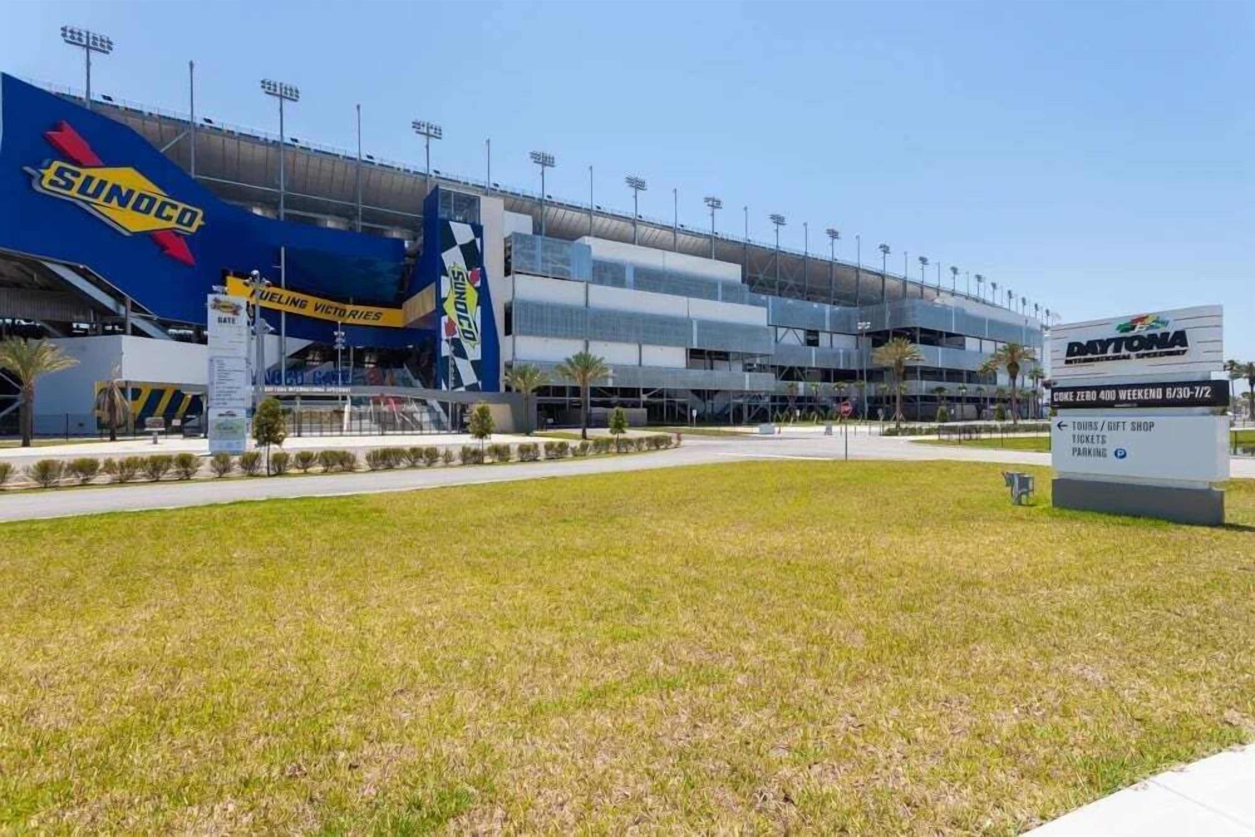 A stadium with a large blue and yellow sign for Sunoco, a modern building with multiple levels, and a sign for Daytona 500 weekend tickets.