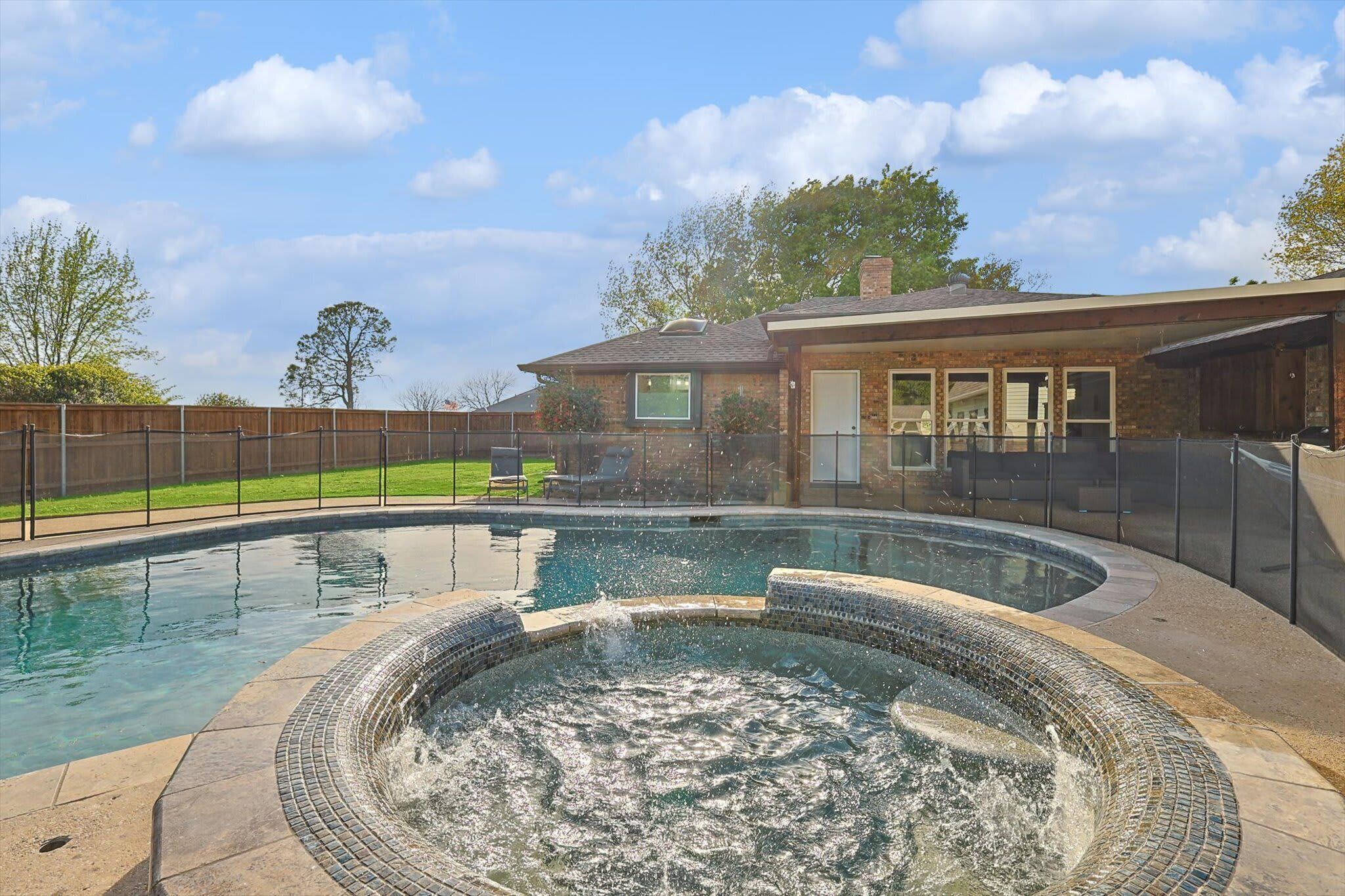 A swimming pool with a hot tub, surrounded by a fence, and a house in the background.