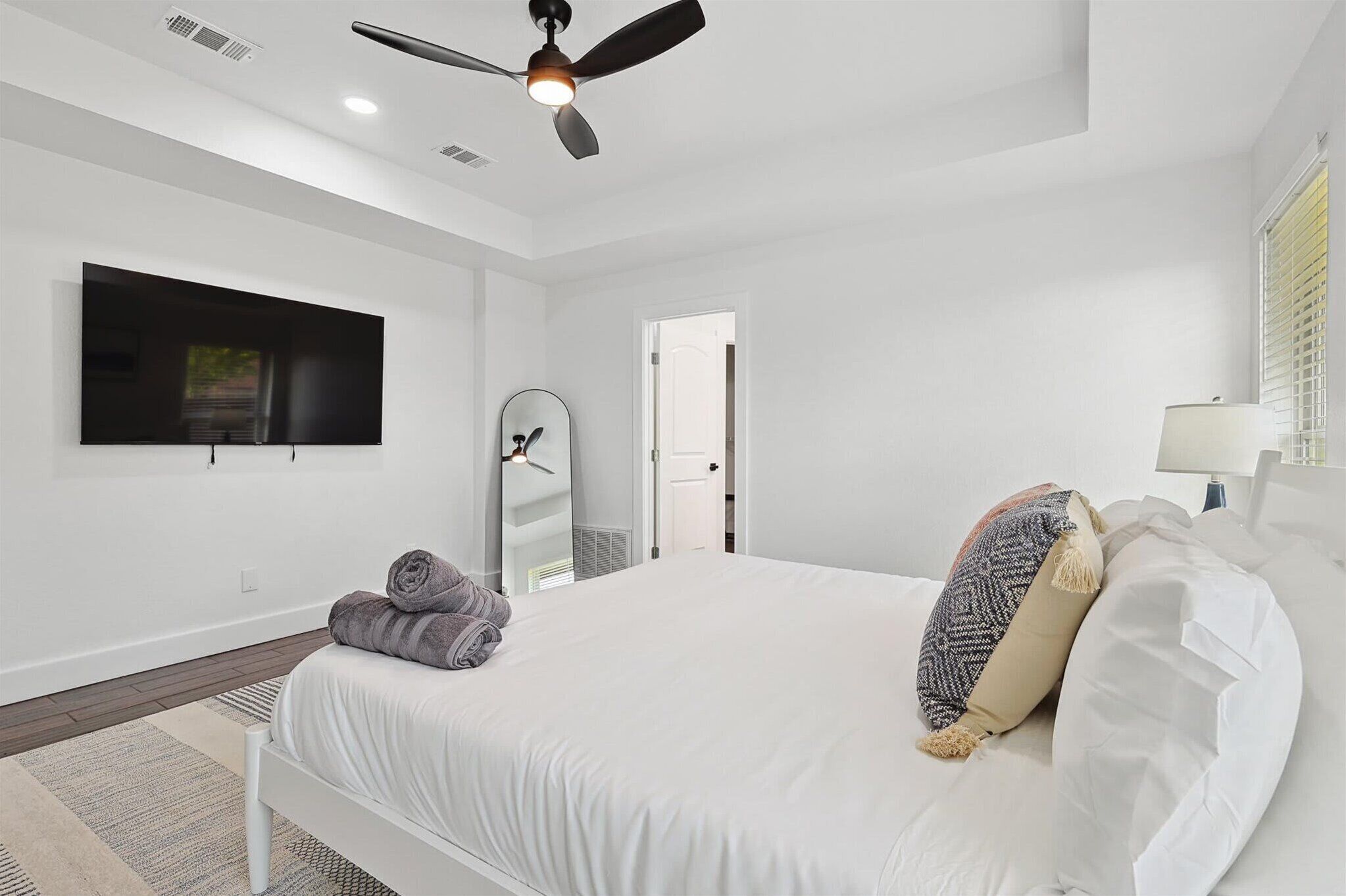 A modern bedroom with a bed, a ceiling fan, a TV, a mirror, and a door.