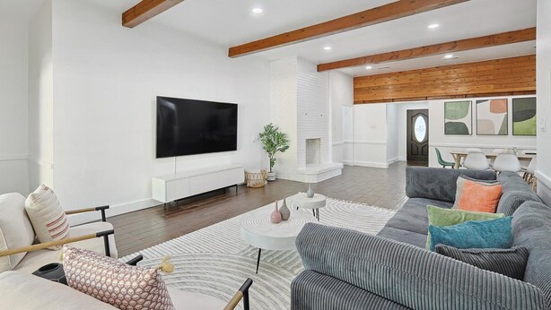 A modern living room with a flat-screen TV, a grey sofa set, a white fireplace, and wooden beams on the ceiling. - Gorgeous 4BR Home With Huge Yard, Pool (Denton)