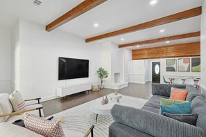 A modern living room with a flat-screen TV, a grey sofa set, a white fireplace, and wooden beams on the ceiling. - Gorgeous 4BR Home With Huge Yard, Pool (Denton)