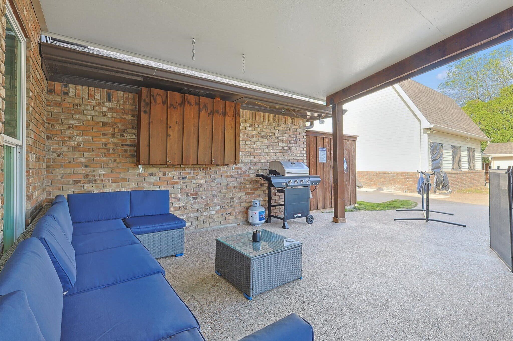 Un espace patio extérieur agrémenté d’un mur en briques, d’une armoire en bois, d’un barbecue, de mobilier d’extérieur bleu et d’une table à plateau en verre.