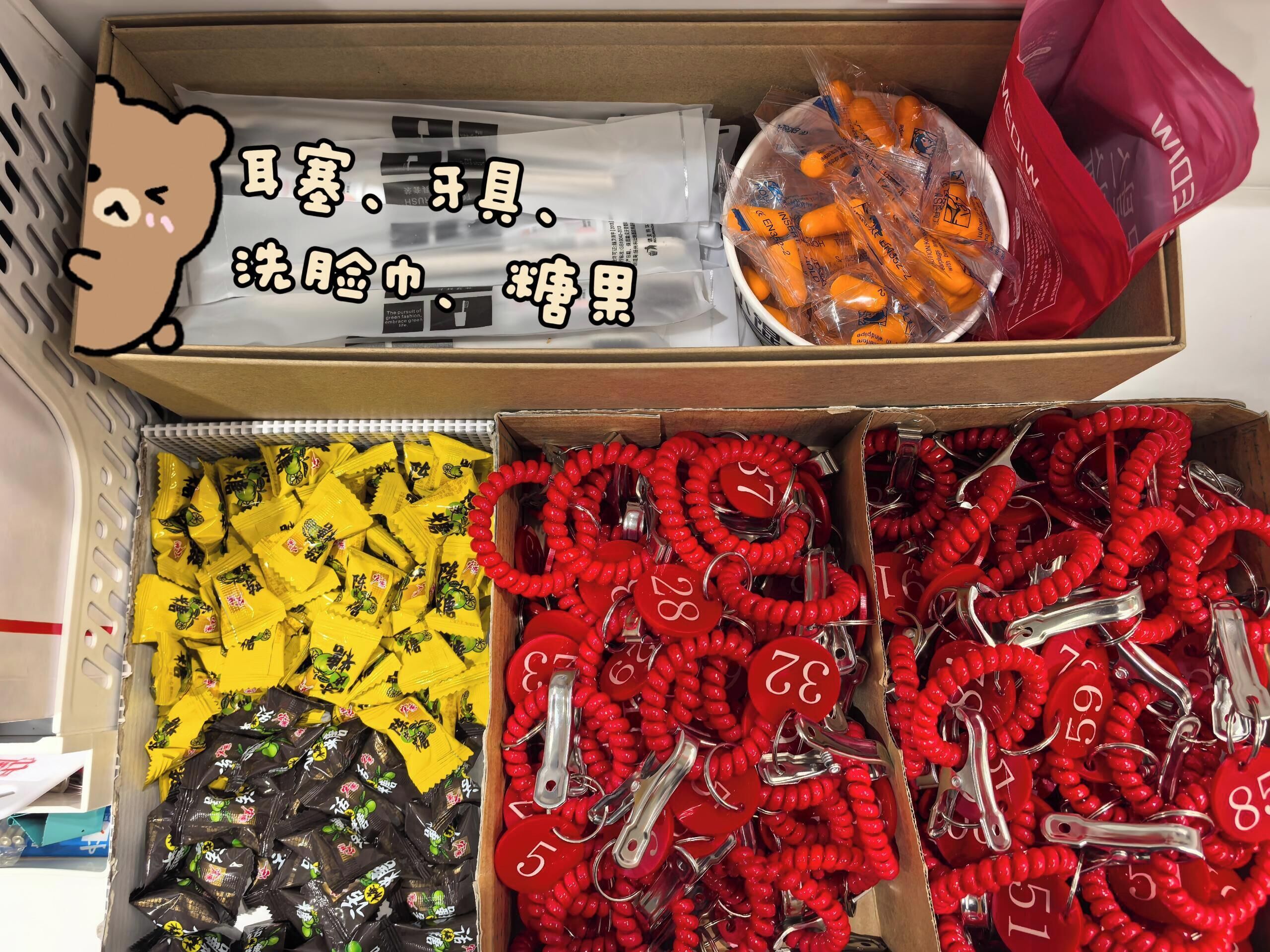En kasse med forskellige ting, herunder snacks i gul og sort emballage, røde nøgleringe med tal, og orange genstande i plastic.