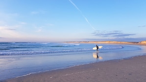 En strand med en person som bär på en surfbräda, en sandstrand och vågor som bryter mot havet.