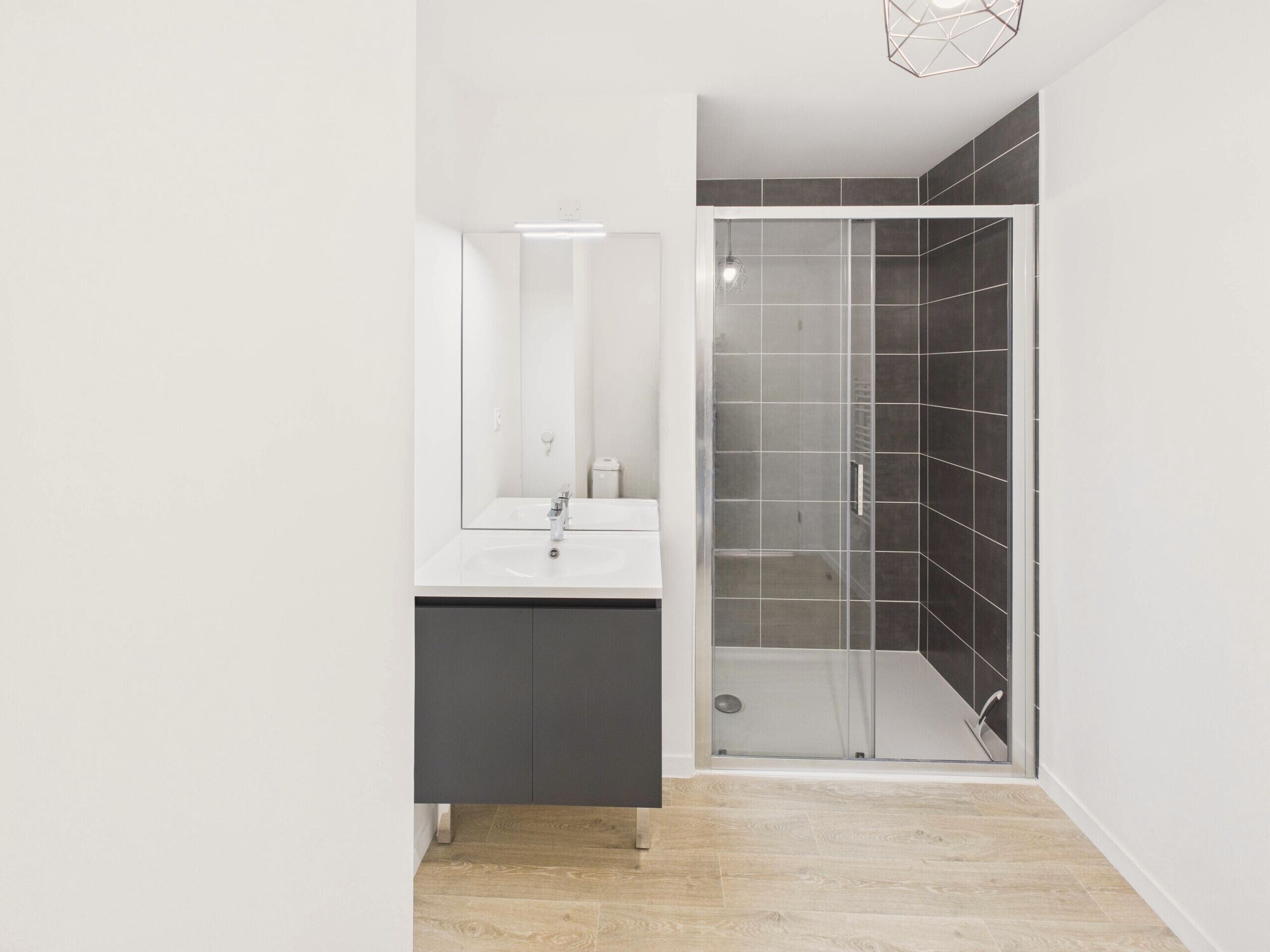 Een moderne badkamer met een wit wastafelmeubel, een douche met donkere tegels en een geometrische lamp.