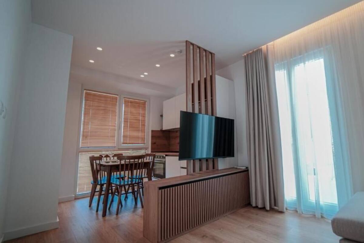 A modern living room with a flat-screen TV, a dining area with wooden chairs, and a sofa.