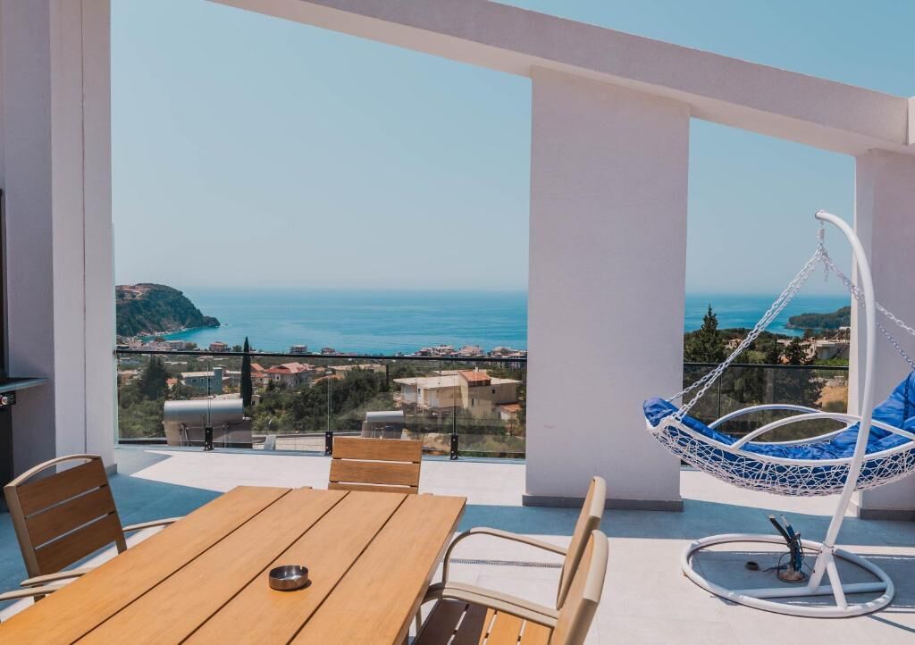 A balcony with a wooden table and chairs, a hanging chair, and a view of the sea and buildings.
