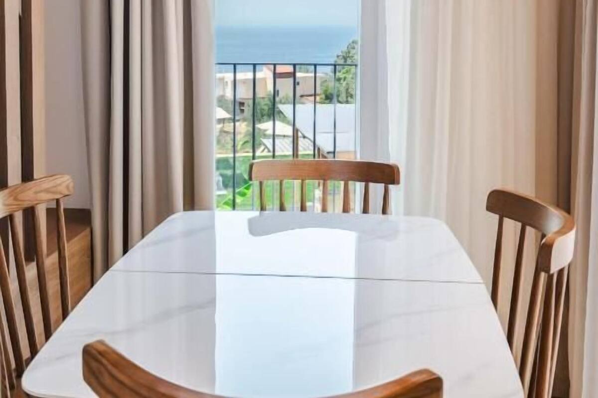 A dining area with a white table and wooden chairs, a woven pendant light, and a view of the sea through large windows with curtains.