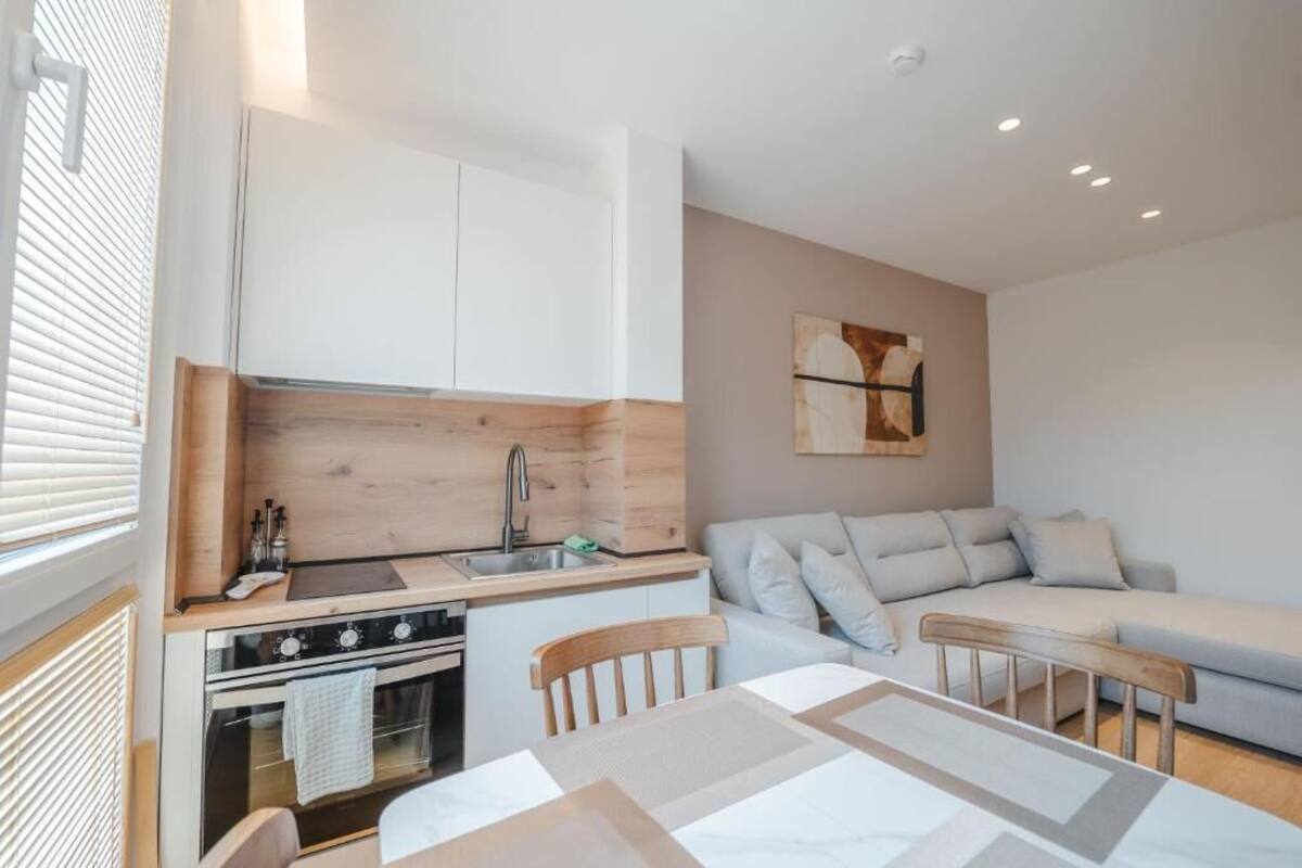 A compact kitchen with a stove, sink, and wooden cabinets. There is a dining table with chairs and a sofa in the living area.